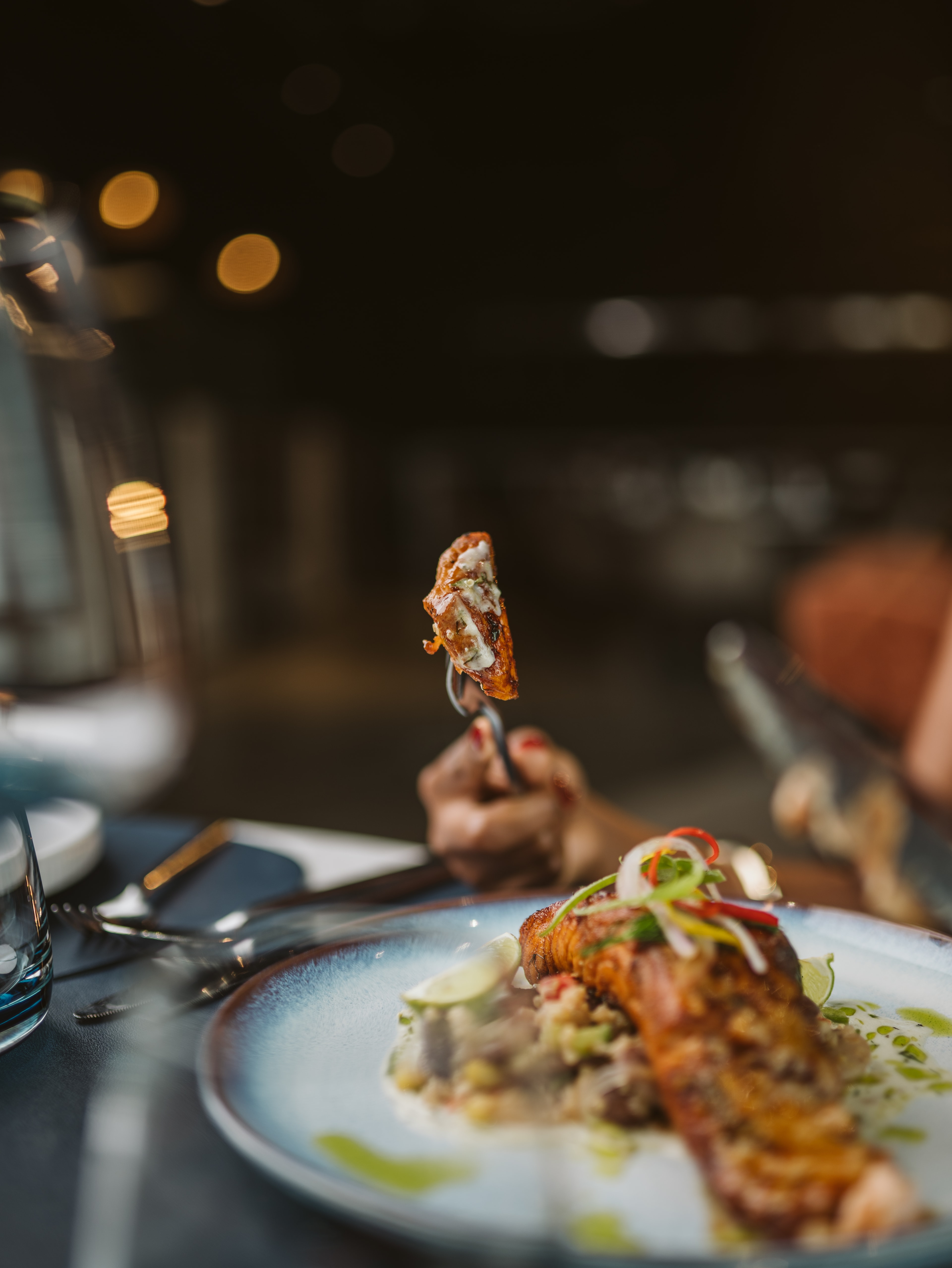 Seared fish fillet with chili and herb oil on a white plate, a diner lifting a bite on a fork under warm restaurant lights